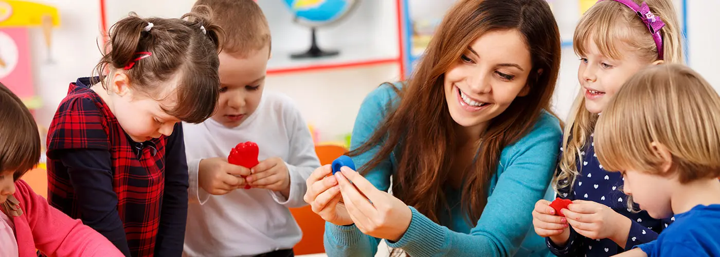 Children stand around their nursery teacher holding playdough molds. The teacher smiles as she shows one mold to a girl.