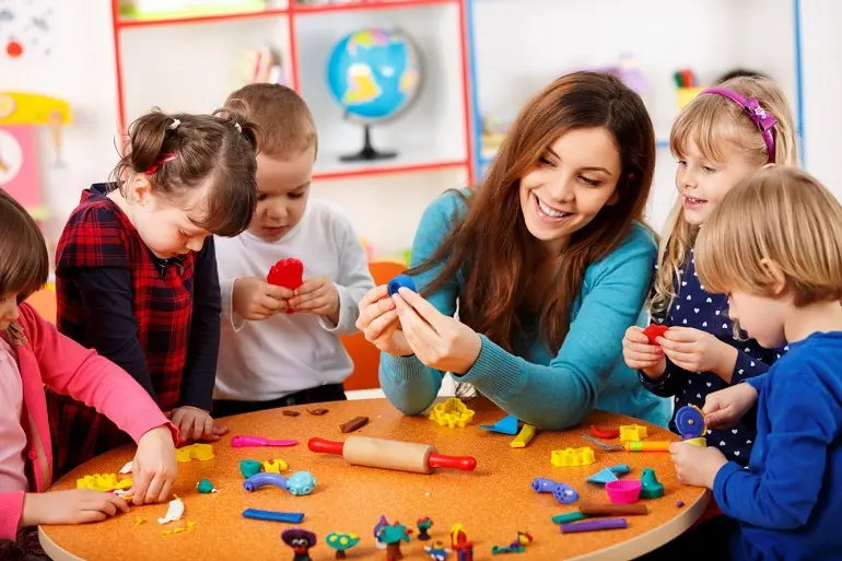 Children stand around a table at nursery with playdough, molds & toy tools. The teacher is showing one mold to a child.