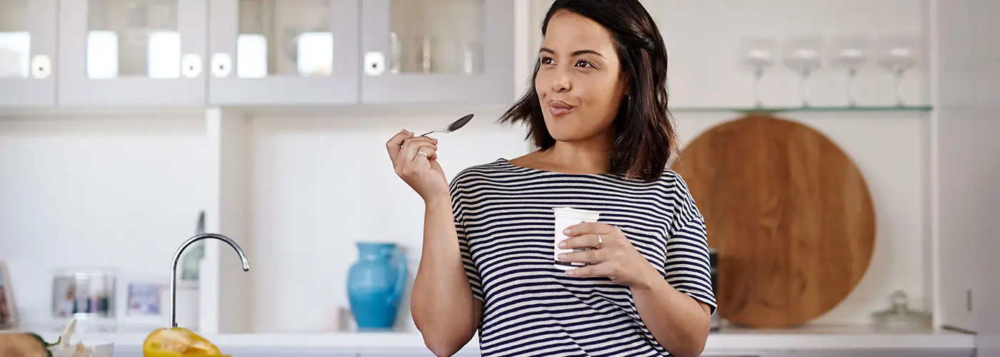 A close up of a woman in her kitchen smiling as she looks off into the distance after taking a mouthful of yogurt.