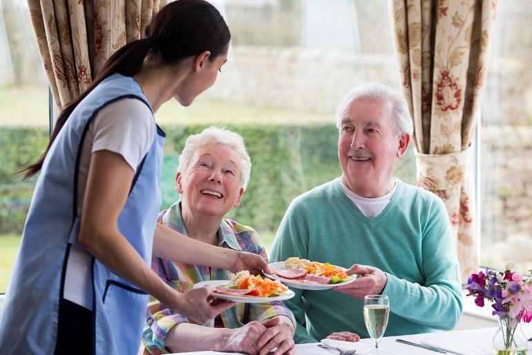 A caretaker hands two plates of salad over to an elderly couple who are sitting at a table & smiling.