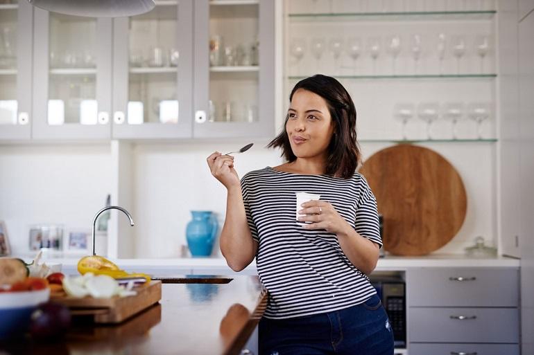 A woman leans against her kitchen island, which displays many vegetables, while she takes a mouthful of yogurt.