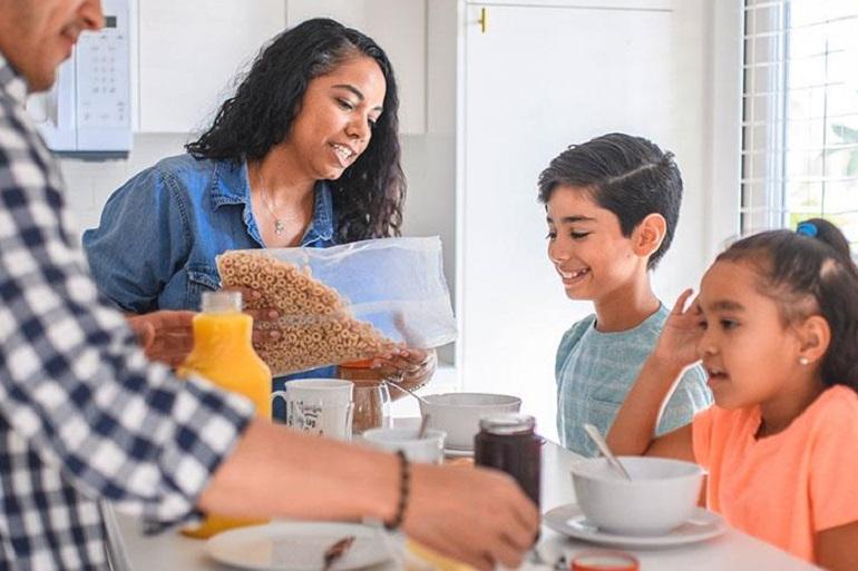 Close-up of a family at a table that's prepared for breakfast. The mother pours cereal into her son's bowl & he smiles.