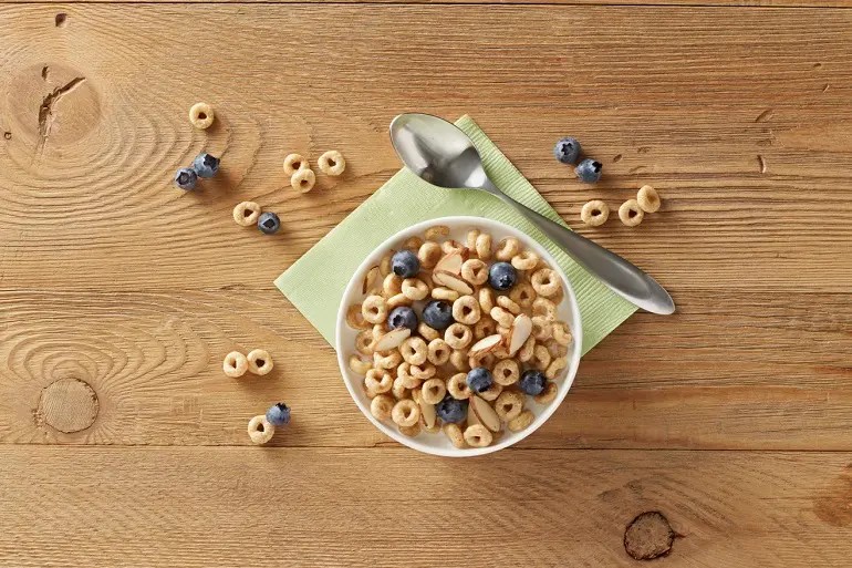 Cereal & blueberries in a white bowl by a metal spoon on a wooden table. Cereal pieces and blueberries are dotted on the table.