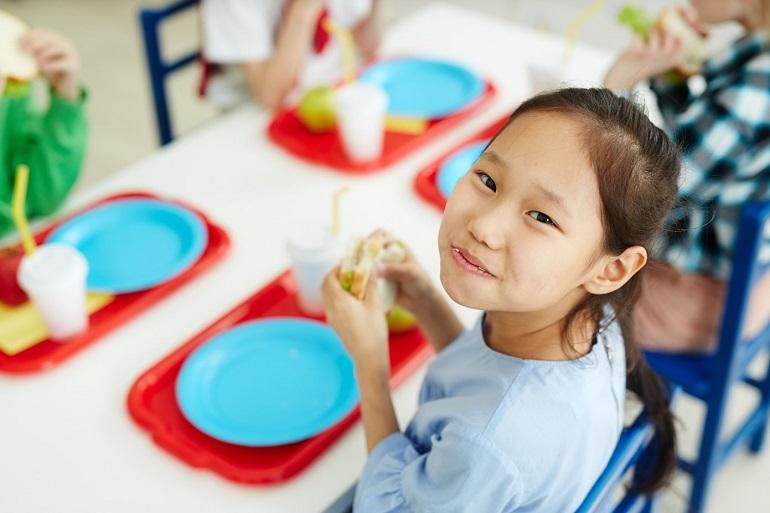 A girl in a blue top sitting at a lunch table looks up toward the camera smiling after taking a bite of her sandwich.