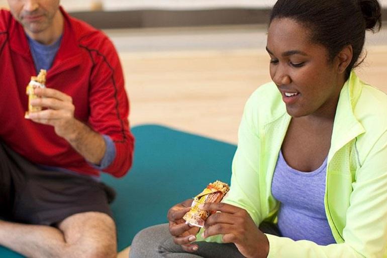 Close-up of a man & woman dressed in workout clothes enjoying cereal bars while sitting on a padded workout mat in a gym.