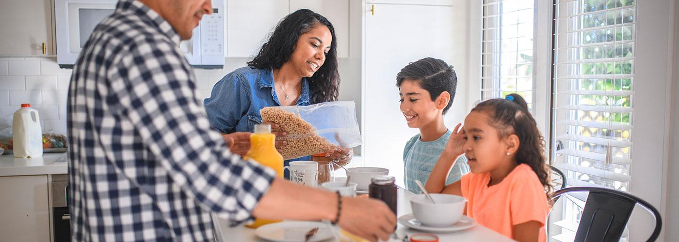 A family get ready to eat breakfast together in their kitchen. The son smiles as his mother pours cereal into his bowl.