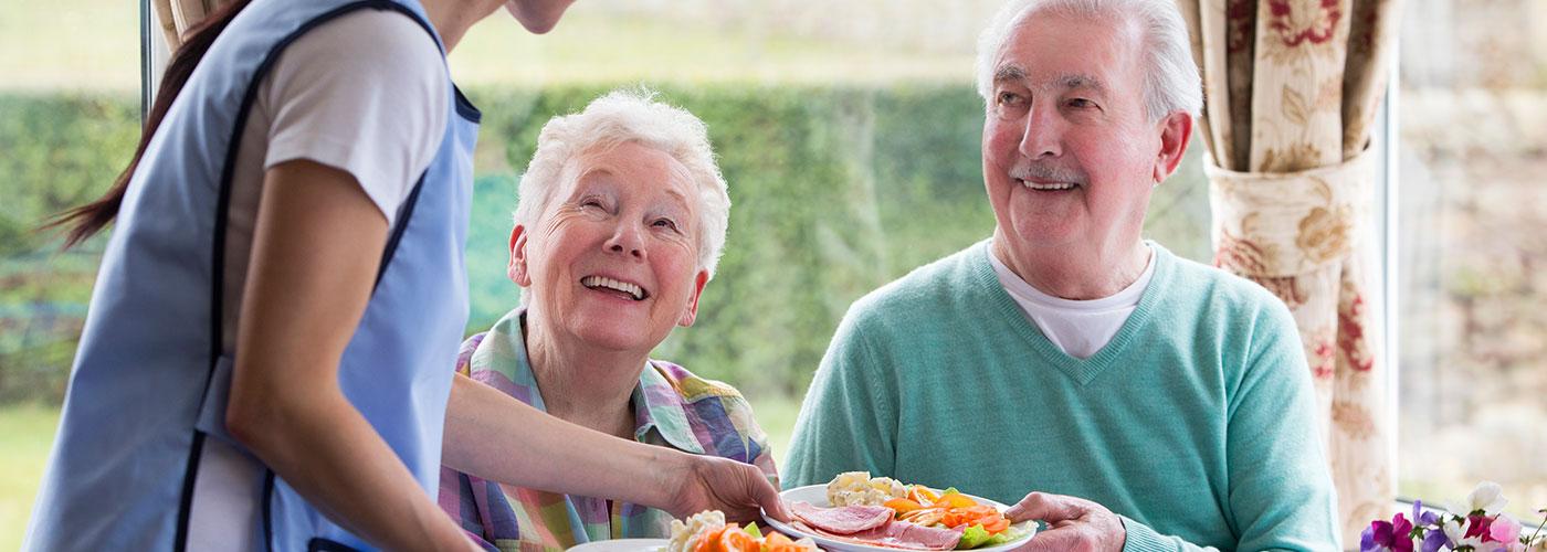 An elderly couple smile up at a caretaker as she hands them each a plate consisting of vegetables and ham.