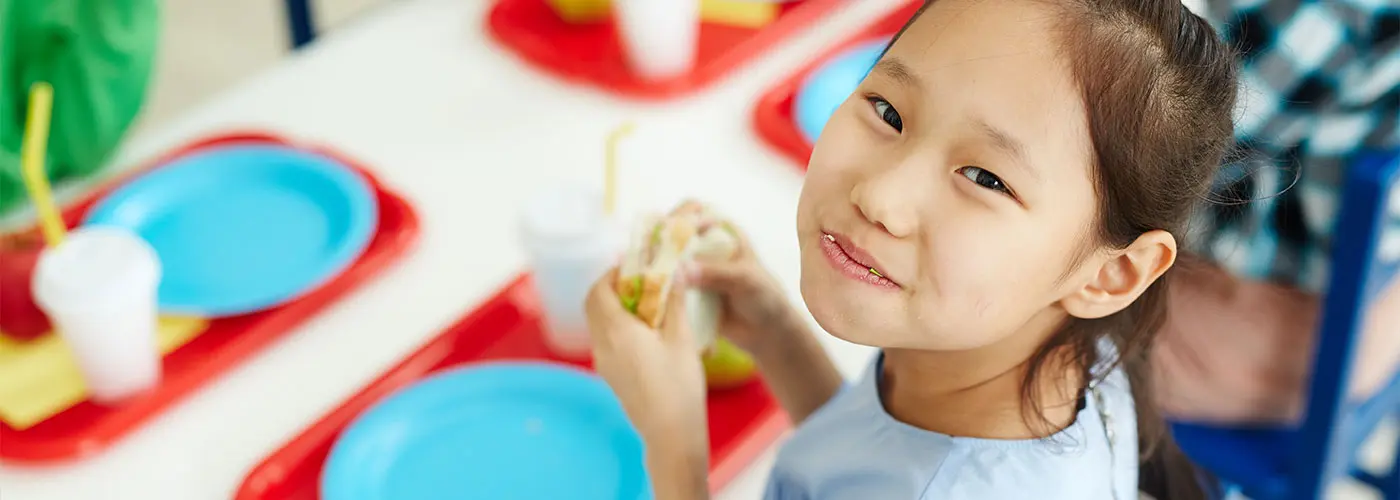 Close-up of a girl sitting at a lunch table looking toward the camera & smiling after taking a bite of her sandwich.