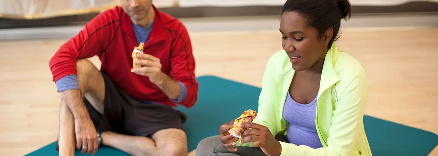 A man & woman wearing workout clothes enjoy cereal bars while sitting on a padded workout mat in a gym.
